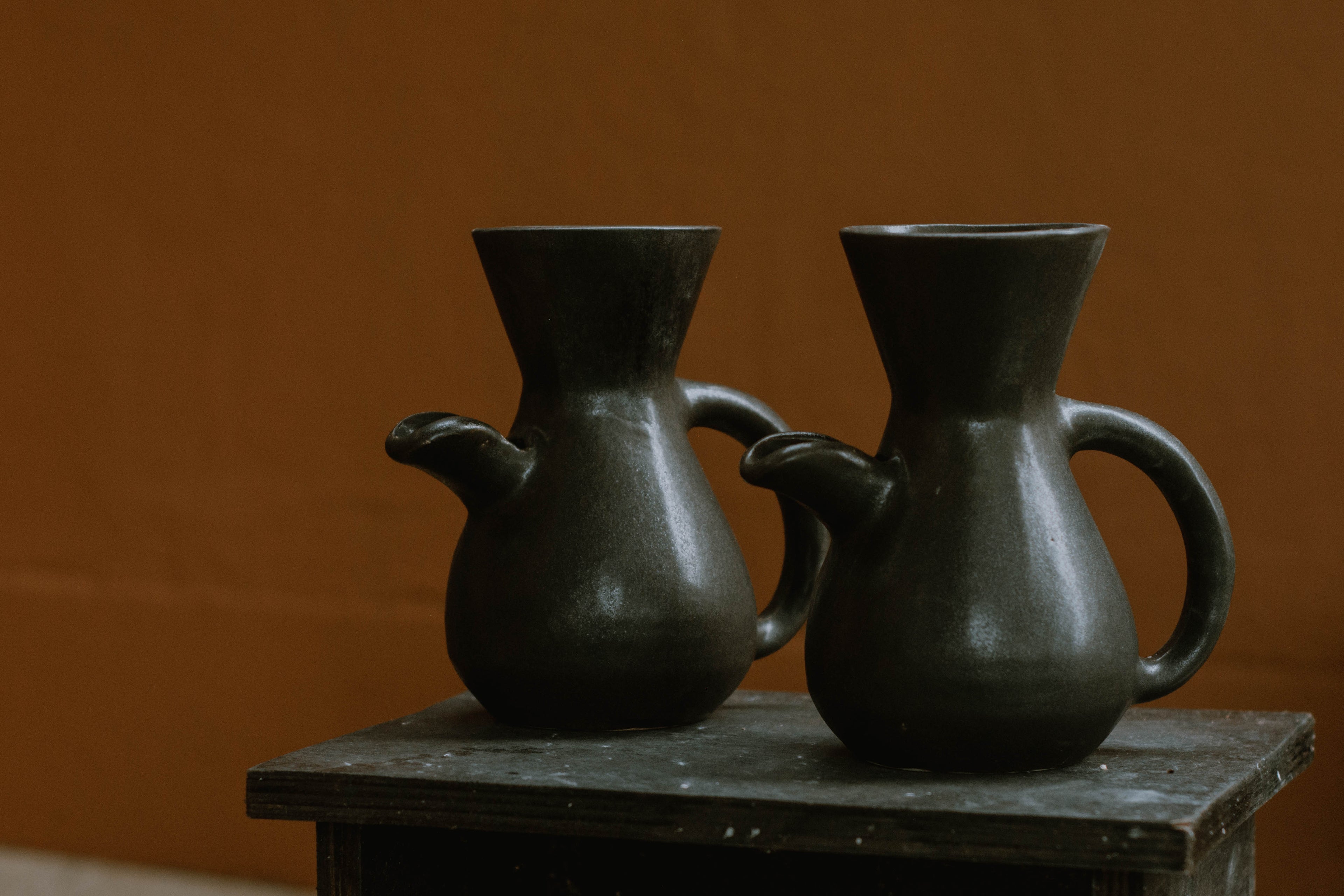 The Kápi Coffee Maker, a handcrafted clay pot from Costa Rica, with a matte black finish. Its elegant, simple design features a flared top, sturdy handle, and a distinctive oxygenation hole for enhancing the coffee's flavor. The artisanal craftsmanship is evident in the pot's textured surface and organic shape, embodying a blend of tradition and functionality in its compact form.