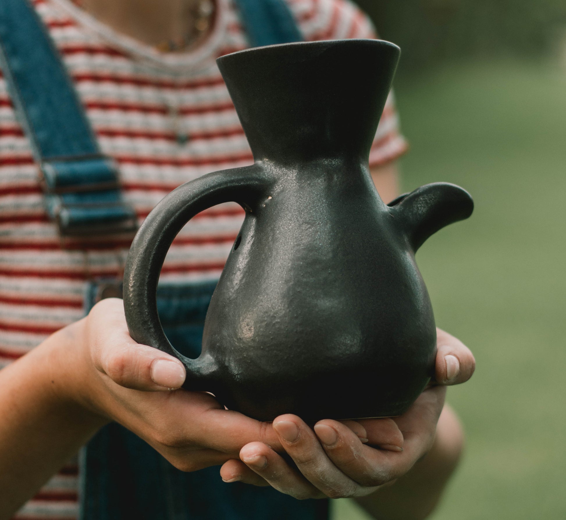 "The Kápi Coffee Maker, a handcrafted clay pot from Costa Rica, with a matte black finish. Its elegant, simple design features a flared top, sturdy handle, and a distinctive oxygenation hole for enhancing the coffee's flavor. The artisanal craftsmanship is evident in the pot's textured surface and organic shape, embodying a blend of tradition and functionality in its compact form."