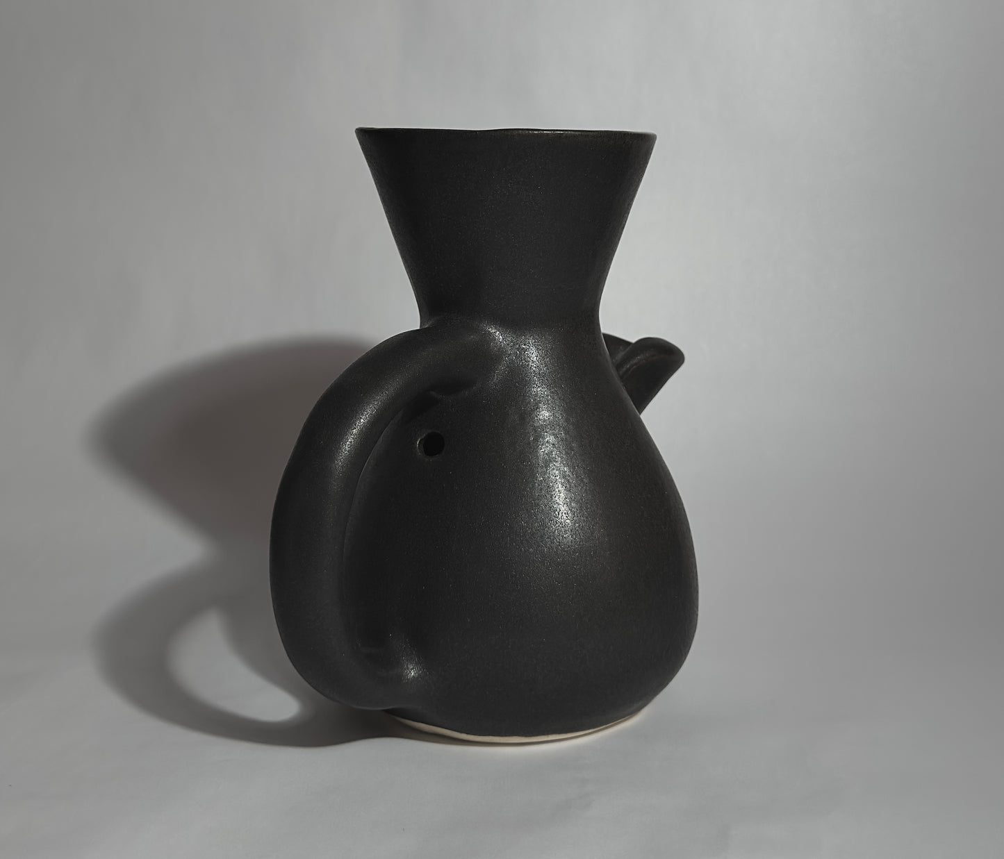 "The Kápi Coffee Maker, a handcrafted clay pot from Costa Rica, with a matte black finish. Its elegant, simple design features a flared top, sturdy handle, and a distinctive oxygenation hole for enhancing the coffee's flavor. The artisanal craftsmanship is evident in the pot's textured surface and organic shape, embodying a blend of tradition and functionality in its compact form."