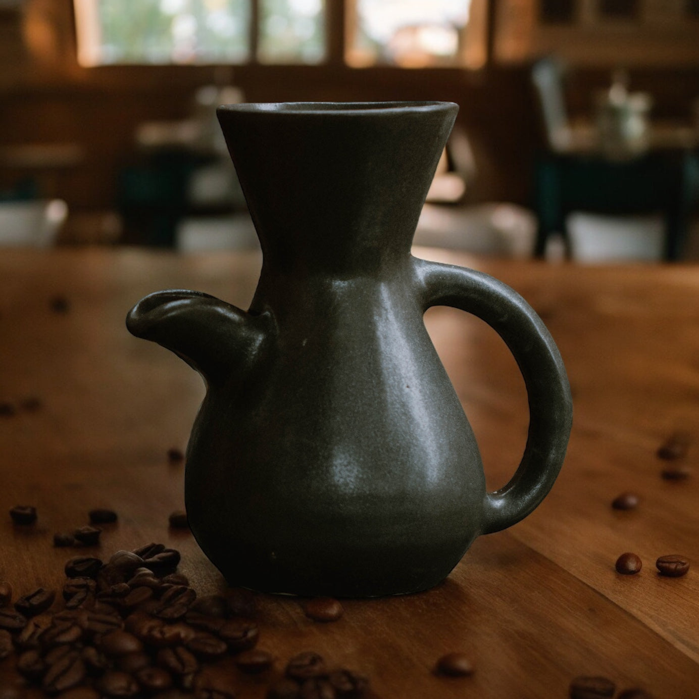 The Kápi Coffee Maker, a handcrafted clay pot from Costa Rica, with a matte black finish. Its elegant, simple design features a flared top, sturdy handle, and a distinctive oxygenation hole for enhancing the coffee's flavor. The artisanal craftsmanship is evident in the pot's textured surface and organic shape, embodying a blend of tradition and functionality in its compact form.