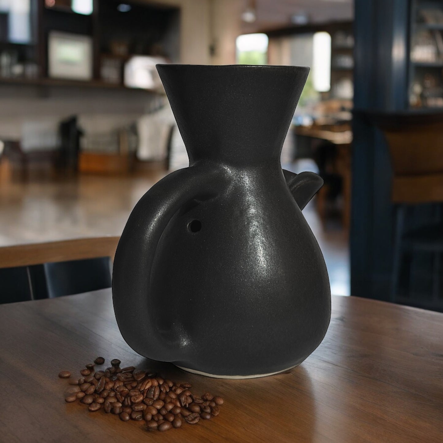 The Kápi Coffee Maker, a handcrafted clay pot from Costa Rica, with a matte black finish. Its elegant, simple design features a flared top, sturdy handle, and a distinctive oxygenation hole for enhancing the coffee's flavor. The artisanal craftsmanship is evident in the pot's textured surface and organic shape, embodying a blend of tradition and functionality in its compact form.